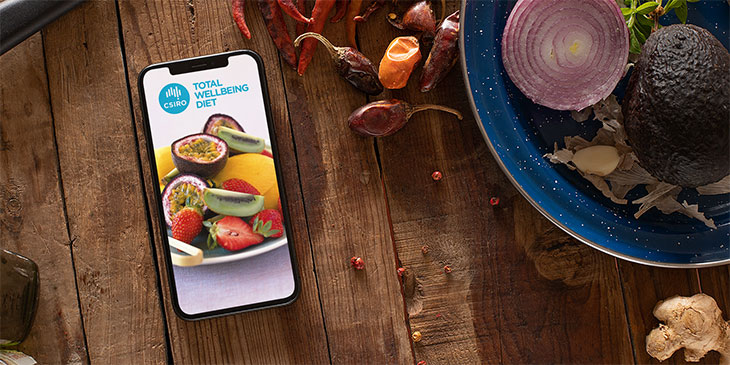 iphone with total wellbeing diet app on rustic table next to recipe ingredients including red onion, avocado and chillies