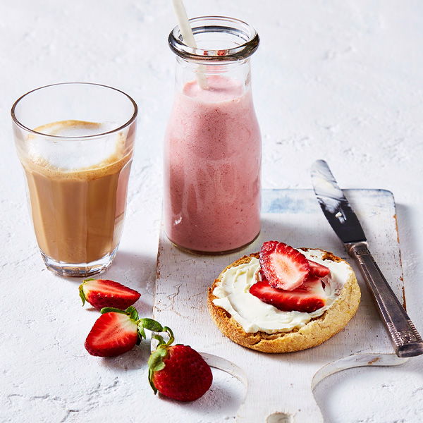 An english muffin with cream cheese and strawberries, next to a healthy strawberry smoothie and coffee,