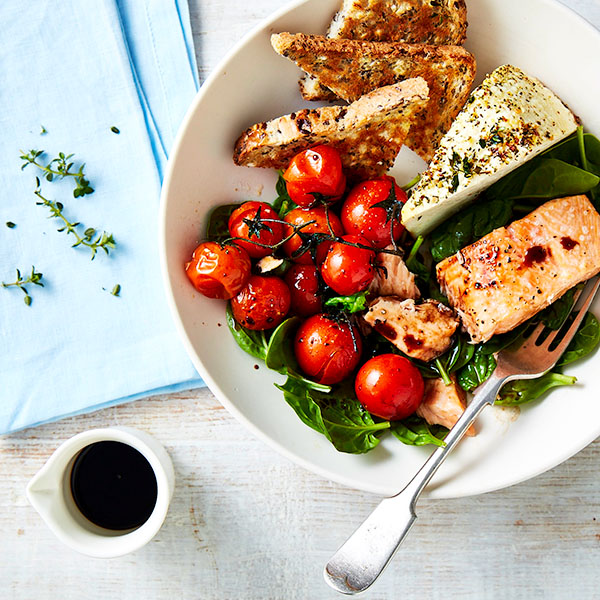 Bowl of salad with salmon, fried bread and feta cheese, baby tomatoes and baby spinach. Next to the plate a small jug of dressing. 