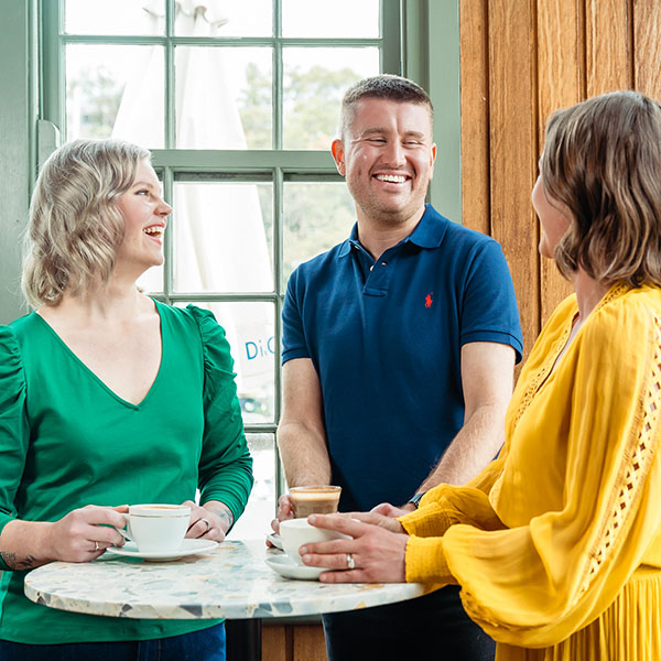 Group shot of members Kayleen (in a green jacket), Gene (in a navy polo shirt) and Jessica (in a yellow, long-sleeved dress) standing at a cafe table drinking coffee