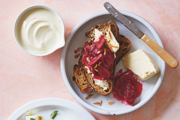 Toasted bread topped with ricotta, stewed rhubarb and flaked almonds, served with a bowl of yoghurt on a pink table