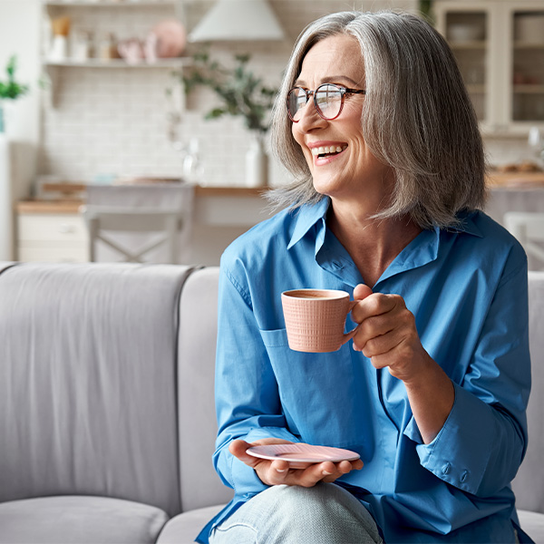 Elderly woman with glasses and a blue shirt sitting on a couch and enjoying a cup of tea